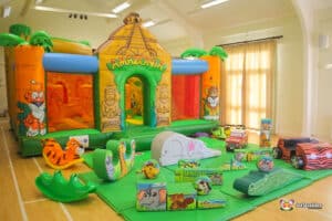 A colorful inflatable jungle-themed bouncy castle with animal designs, surrounded by soft play toys, all indoors.