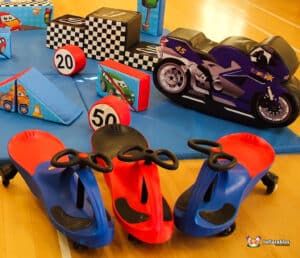 A colorful play area featuring red and blue ride-on toys, inflatable traffic signs, and toy cars on a blue mat with race-themed decor.