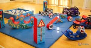 A colorful indoor play area featuring soft play traffic-themed obstacles, a ball pit, and ride-on toy cars for children.