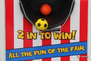 A colorful carnival game with a black target holding a yellow soccer ball and an orange bouncy ball, labeled "2 in to win!"