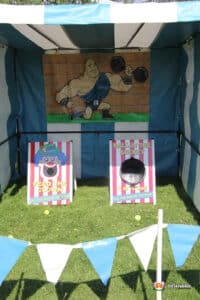 A colourful carnival side stall game booth featuring a muscular cartoon character, with two games: "Feed the Clown" and "Ball in the Bucket."