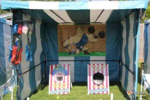 A colourful carnival side stall featuring games like "Feed the Clown" and "Ball in the Bucket," with playful decorations and bright signage.