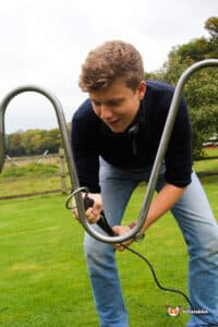 A person kneels on grass, holding an air pump attached to a large metal frame, preparing to inflate an object outdoors.