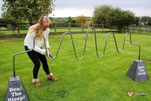 A person navigates a wavy metal challenge game in a grassy area, aiming to avoid touching the metal frame, with signs reading "Beat The Bleep!"
