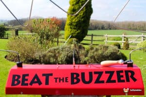 A vibrant red game console labeled "BEAT THE BUZZER" set in a lush garden with green fields and hedges in the background.