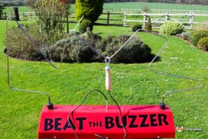 A red "Beat the Buzzer" game with metal wire loops and a wooden handle, set on a grassy field with bushes and trees in the background.