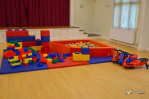 Colorful soft building blocks and a ball pit are set up in a spacious play area, accompanied by toy ride-on cars.