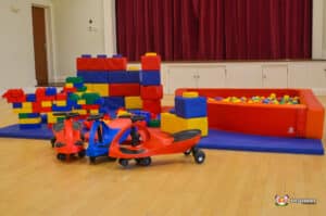 A colorful indoor play area for kids featuring building blocks, a ball pit, and ride-on toys against a red curtain backdrop.