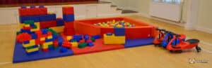 A colorful children's play area featuring a ball pit, soft blocks, building blocks, and ride-on toys on a wooden floor.