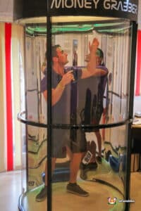 A man in a purple shirt stands inside a glass box, resembling a cash grabber machine filled with bills and tokens.