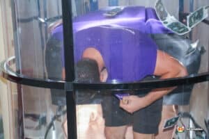 Inside a glass box, a man in a purple shirt reaches for cash, mimicking a cash grabber game filled with money.