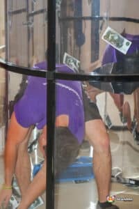 A man wearing a purple shirt is inside a glass box, simulating a cash grabber, surrounded by flying bills.