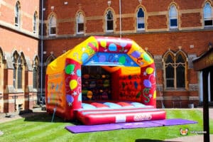 A colorful inflatable bounce house set in a sunny courtyard, surrounded by brick buildings featuring decorative windows.