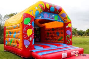 A colorful inflatable bounce house with bright balloons and stars, set on a grassy area under a cloudy sky.
