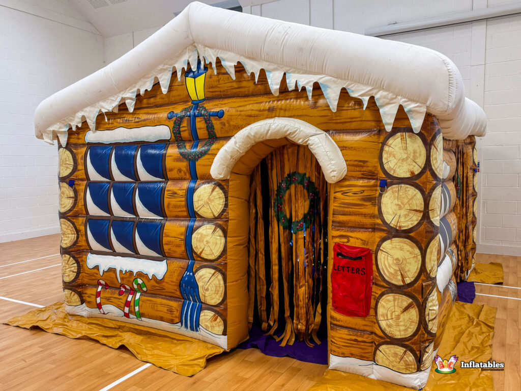 Angled exterior view of the inflatable Christmas Grotto cabin, highlighting the arched doorway with a green wreath and the log-style construction, set up in a large indoor hall