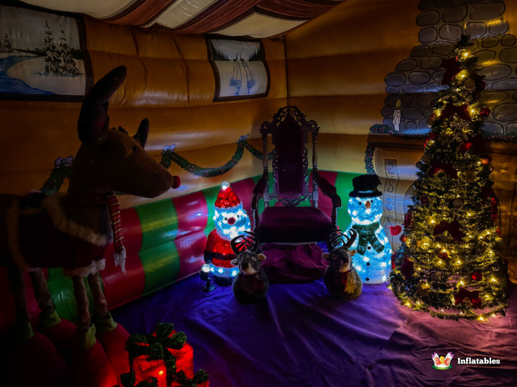 Darker interior view of the grotto, showing the Santa's throne surrounded by festive decorations, including a plush reindeer prop, glowing figures, and a lit Christmas tree