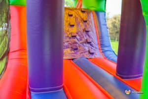 Brightly colored inflatable slide featuring purple pillars, red and blue surfaces, and a climbing wall with grips. Fun outdoor play equipment.