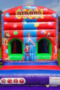 A colorful circus-themed inflatable bounce house featuring playful animals, balloons, and a vibrant design, set against a sunny blue sky.