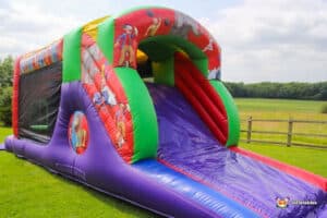 A colorful inflatable bounce house slide featuring clown graphics, set on green grass with fields in the background.