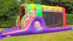 A colorful inflatable bounce house with a slide, featuring playful animal designs, set on a grassy area surrounded by trees.