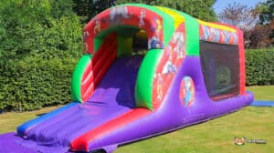 Colorful inflatable slide with a bouncy entrance, set on a grassy area surrounded by lush greenery. Fun and playful design for kids.