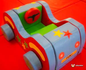 A toy car featuring a red and blue seat alongside a green and blue seat, set in a circus-themed soft play area.