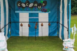 A playful carnival game stall called "Coconut Shy," featuring coconuts on sticks, with a colorful striped background and bags of coconuts nearby.