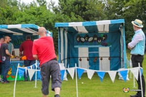 A vibrant outdoor coconut shy side stall with game stalls including "Coconut Shy," attendees engaged in throwing balls, and festive decorations.