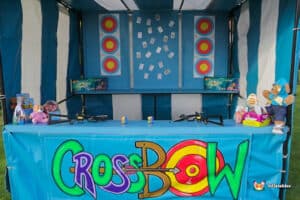 A colourful crossbow shootout side stall booth labeled "CrossBow" featuring targets, toy weapons, and plush prizes under a blue and white canopy.