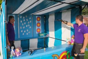 A carnival game crossbow shootout side stall with colourful targets and prizes. A player aims a toy gun, while another person stands nearby, watching.