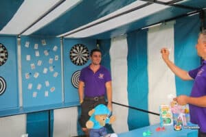 A fairground side stall features two men in purple shirts, darts boards, and a table with toys and prizes. Colourful decorations enhance the festive atmosphere.