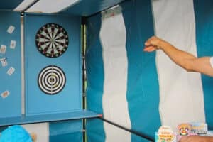 A player throws a dart at a target in a blue and white striped booth, with playing cards displayed on one wall.