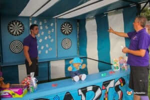 A colourful fairground darts side stall booth features dartboards, plush toys, and games, with two attendants engaged in a discussion.