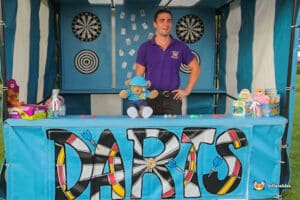 A colorful dart game booth featuring a dartboard, plush toys, and assorted prizes, with a vendor in a purple shirt.