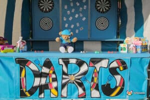 A colorful dart game booth featuring a large "DARTS" sign, dartboards, and a teddy bear mascot sitting at the counter.