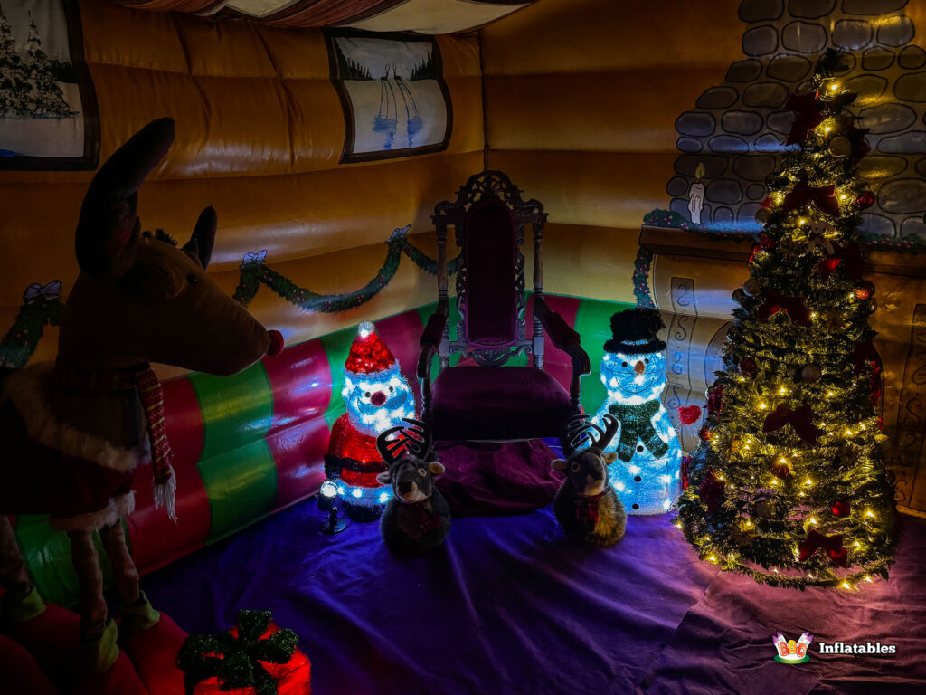 Interior shot of the grotto, featuring a red Santa's throne flanked by a light-up Santa and Snowman, a fully decorated, glowing Christmas tree, and two small plush reindeer.