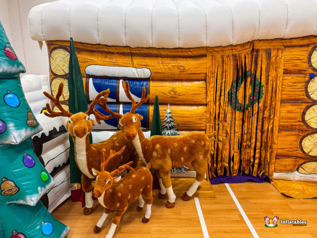 Exterior front view of the inflatable grotto, showing the snow-covered log cabin facade, with three plush reindeer figures and a large inflatable Christmas tree to the side.