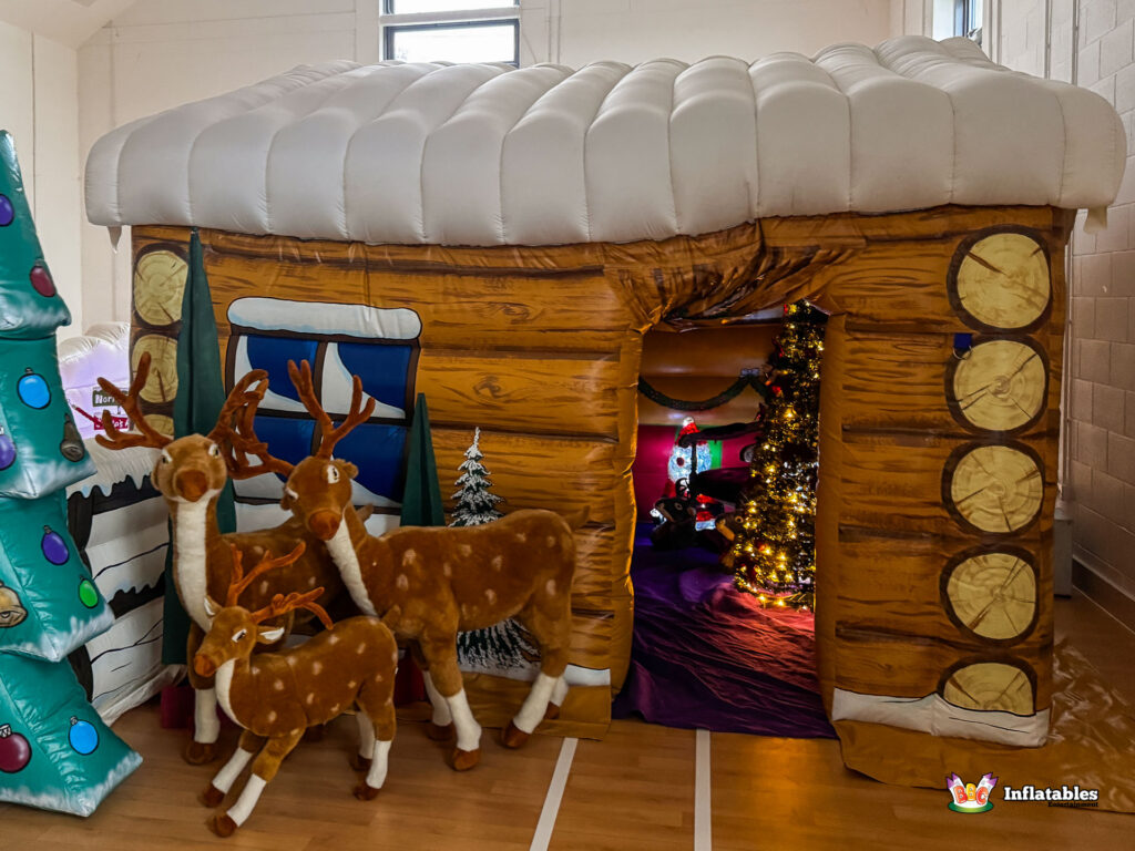 Exterior angled view of the grotto showing the entrance, which reveals the brightly lit interior tree. Three plush reindeer stand guard at the doorway, with an inflatable Christmas tree next to a painted window.