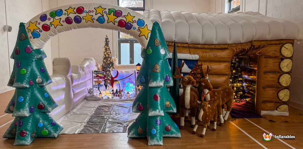 Panoramic exterior view of the Deluxe Christmas Grotto. The log cabin, a snow-covered tunnel archway decorated with ornaments and stars, and a path lined with lights, trees, and reindeer are all visible.