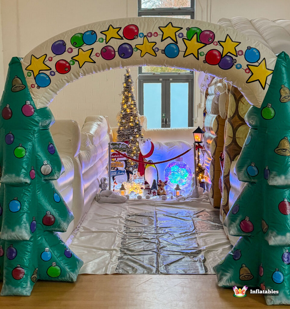 View through the inflatable archway of the Deluxe Grotto. The path is decorated with a white snow sheet, an illuminated Christmas tree, a light-up polar bear, penguin figures, and a decorative signpost pointing to the North Pole.