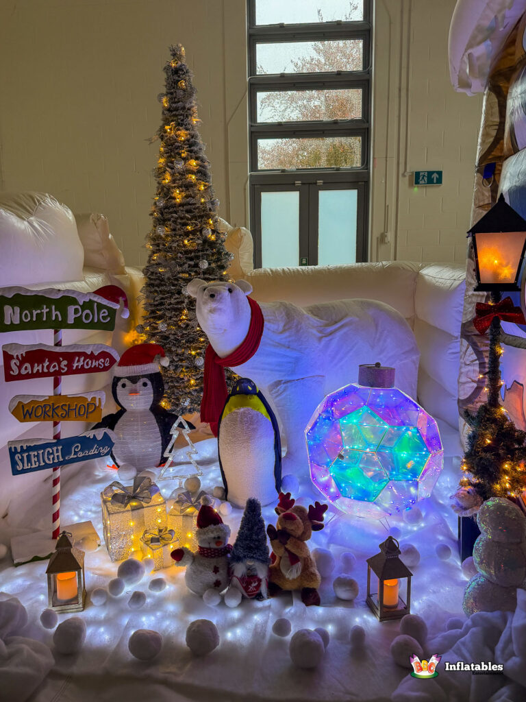 Close-up of the winter scene display area outside the grotto, featuring a tall, lighted Christmas tree, a polar bear, penguins, a North Pole sign, and a large color-changing light-up ornament.