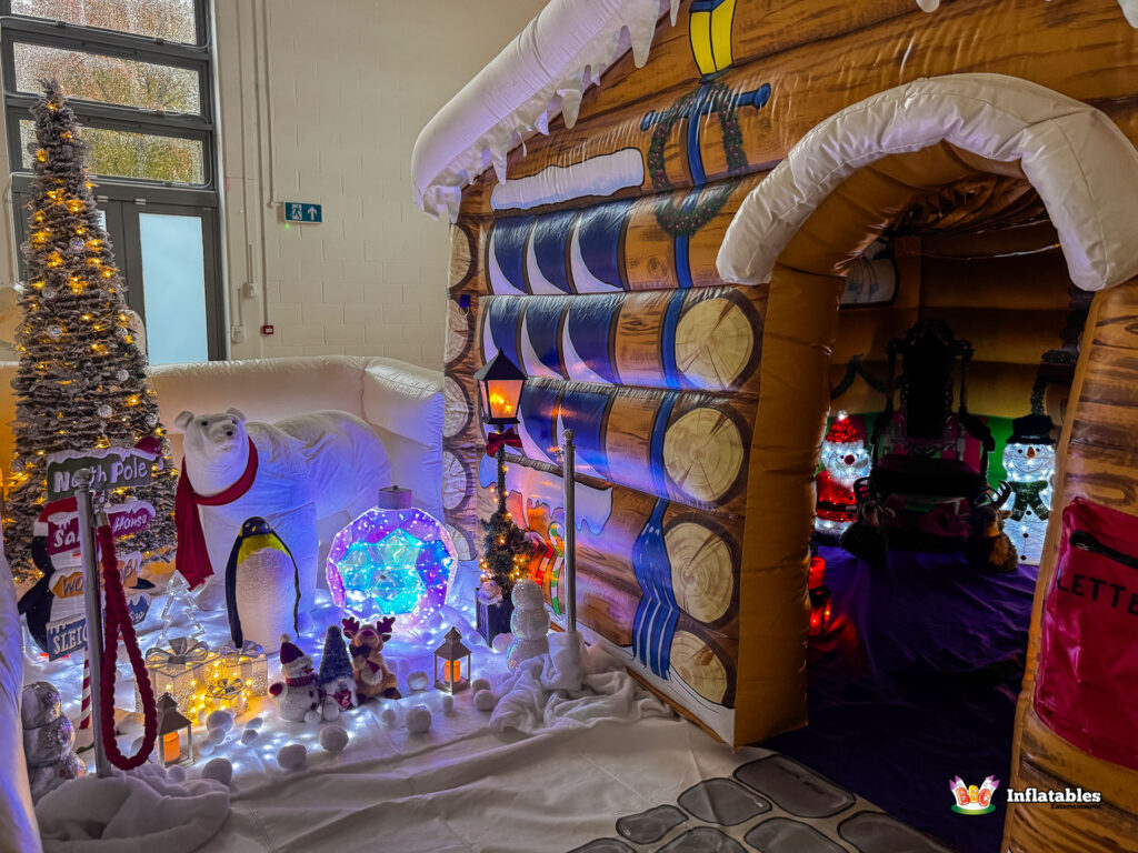 View combining the exterior of the log cabin and the adjacent snow scene display. The cabin entrance is visible on the right, and the winter scene features the North Pole sign, a polar bear, and a glowing ornament.
