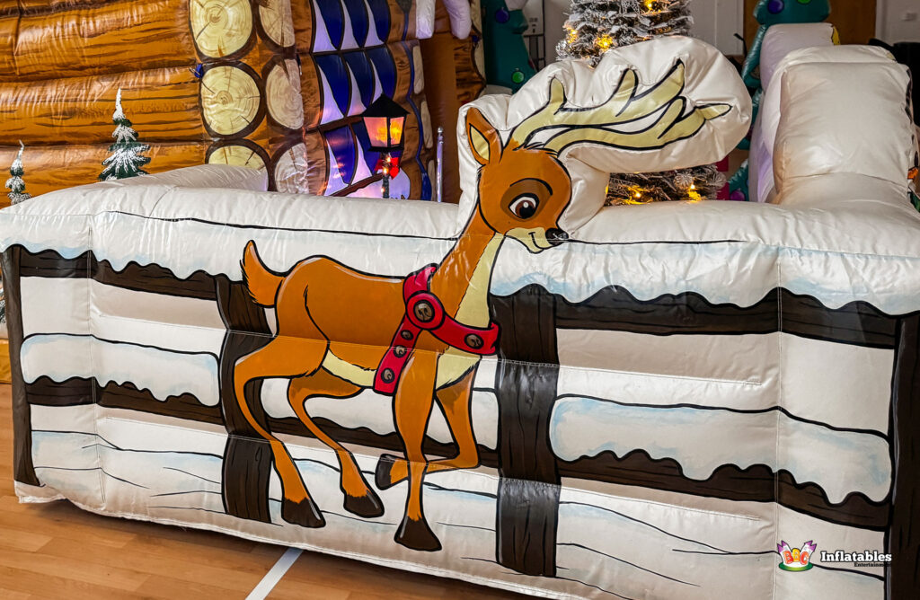 Close-up of the inflatable fence barrier section, featuring a cartoon illustration of a reindeer with white antlers jumping over a snow-covered wooden fence, with the log cabin grotto visible behind it.