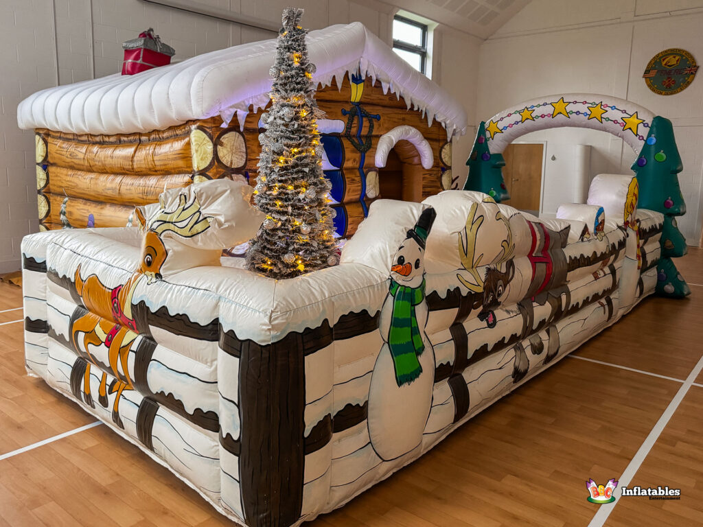 Angled view of the large inflatable barrier, which wraps around the log cabin grotto, decorated with illustrations of snowmen and reindeer. A tall, thin lighted Christmas tree stands on the barrier.