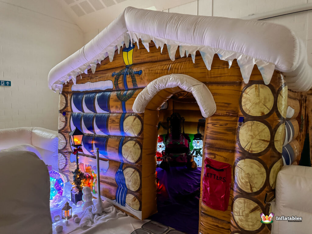 Angled view of the inflatable grotto entrance, with the wood-log design visible. The brightly lit interior decorations are seen through the arched doorway, and the exterior snow scene setup is visible on the left.