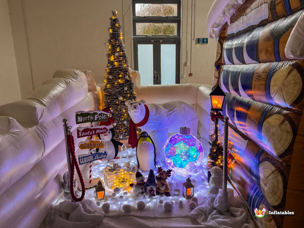 Wide view of the lighted winter wonderland display set up in front of the grotto, including a tall Christmas tree, a polar bear, a penguin, a signpost to the North Pole, and small glowing lanterns.