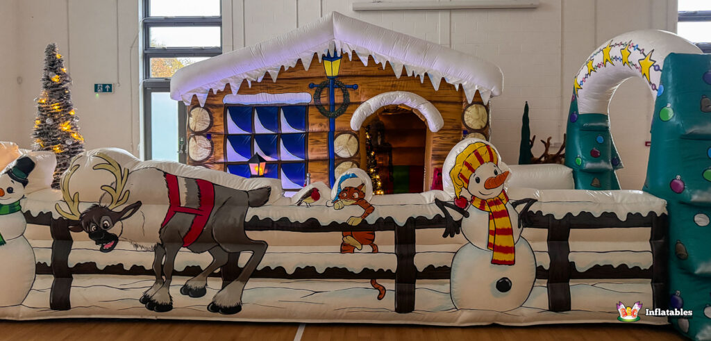 Long frontal view of the inflatable fence barrier and the log cabin grotto backdrop. The fence art shows a snowman with a striped scarf, a reindeer, and a cartoon cat.