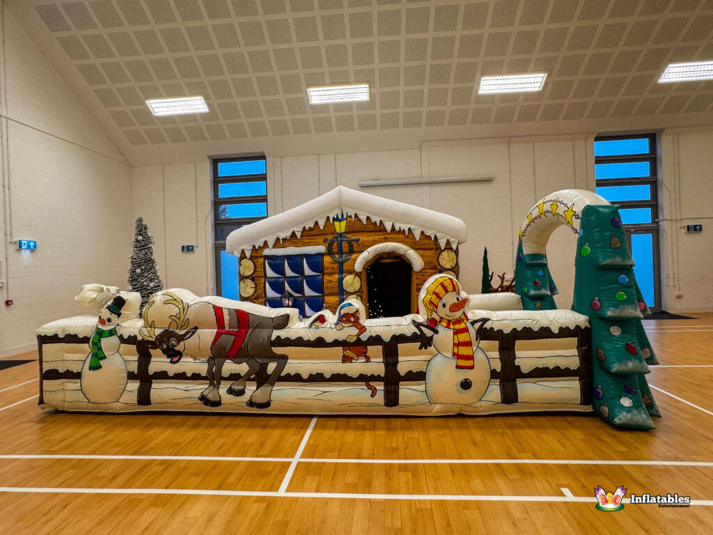 Full view of the combined inflatable attraction, showing the log cabin grotto, the long snowy fence obstacle barrier, and the inflatable Christmas tree archway.