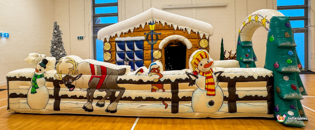 Wide frontal view of the long inflatable fence barrier, set up in front of the log cabin grotto, featuring cartoon artwork of snowmen, reindeer, and a small elf peering over the top.