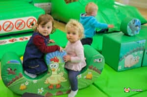 Two young children happily sitting on a green inflatable toy at a Deluxe Farm Soft Play area.